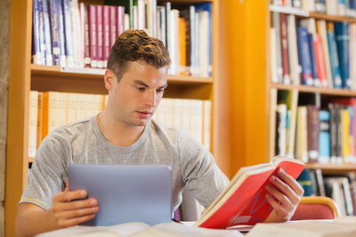Attractive smiling student using tablet and holding book in library